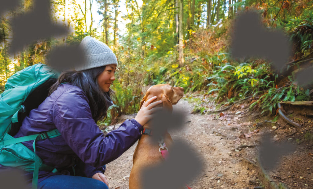 Eine Frau mit Wanderausrüstung, darunter ein Rucksack und eine Mütze, hockt im Wald und streichelt einen Hund. Teile des Bildes sind durch schwarze, unregelmäßige Bereiche verdeckt, die leere oder blinde Stellen symbolisieren.