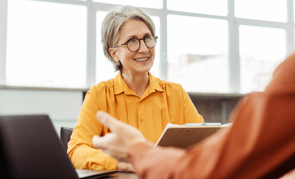 Ältere Frau mit Brille und grauem Haar, die in einem modernen Büro einem Gesprächspartner zuhört